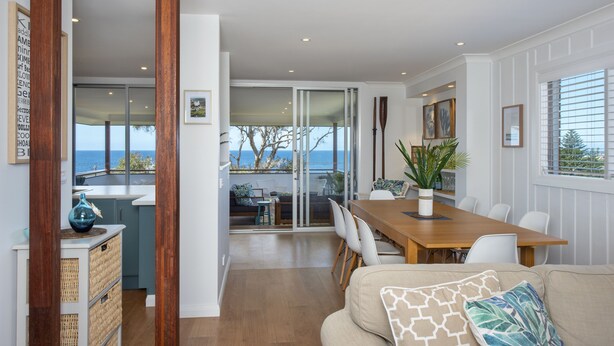 A modern interior with a wooden dining table, white chairs, a beige sofa, and a view of the sea through large windows. - Seascape Kiama: Ocean Views, Family Fun, Pet-Friendly Perfection managed by HAUS (Kiama)