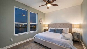 A bedroom with a large bed, two windows with a view, a ceiling fan, and a clock on the wall. - Bayfront Home with Scenic Views & Decks (Galveston)