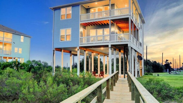 A multi-story waterfront house with a wooden deck leading to the entrance. - Bayfront Home with Scenic Views & Decks (Galveston)