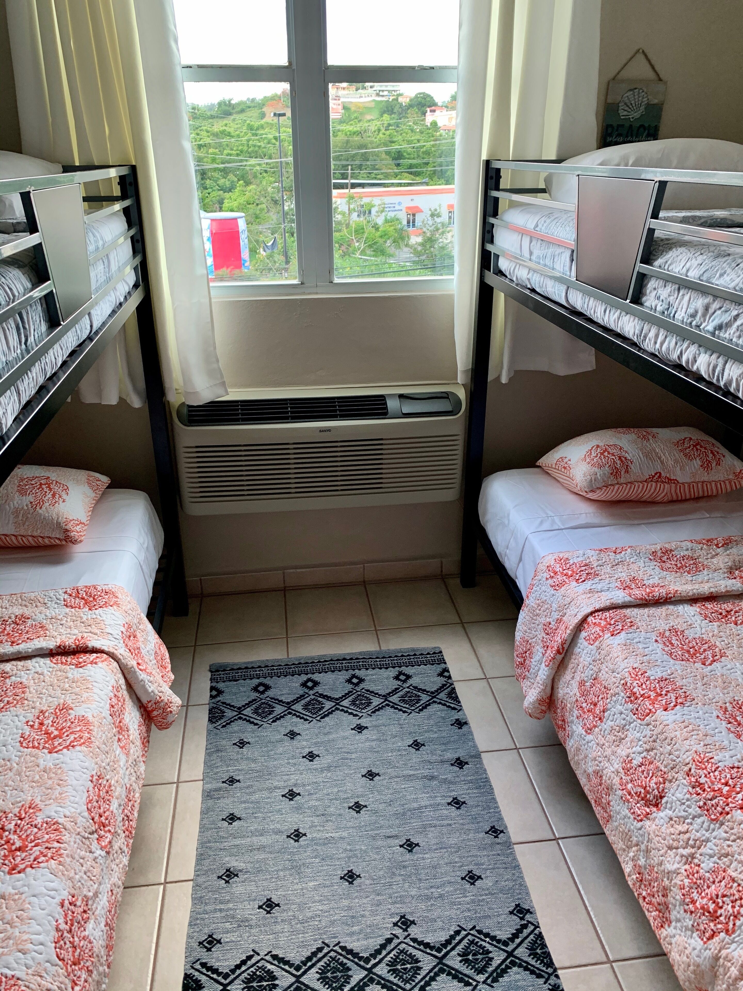 A dormitory room with bunk beds, a window with a view of greenery, a radiator, and a patterned rug.