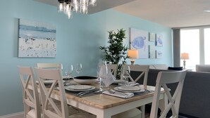 Un comedor con una mesa de madera dispuesta para cuatro personas, rodeada de sillas blancas. Seis lámparas colgantes iluminan la mesa.