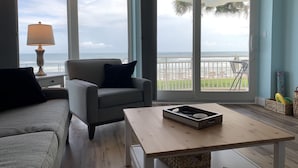 Sala de estar con sofá, sillón y una mesa con bandeja. Amplias ventanas ofrecen vista al océano y un balcón con sillas.