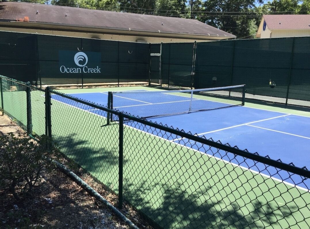 A tennis court with a net, surrounded by a chain-link fence, and a building with the name "Ocean Creek" in the background.