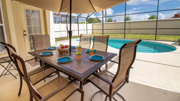 An outdoor patio with a table set for four, surrounded by chairs, and an umbrella providing shade. - Large 4 Bedroom Villa. 3 Miles to Disney, Private Pool, Games Room (Kissimmee)