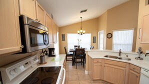 A kitchen with wooden cabinets, a stainless steel microwave, a stove, and a dining area with a chandelier. - Large 4 Bedroom Villa. 3 Miles to Disney, Private Pool, Games Room (Kissimmee)