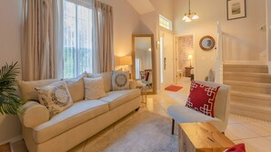 A spacious living room with a beige sofa, a wooden coffee table, and a red and white patterned armchair. A staircase leads to an upper floor, and a dining area is visible in the background. - Reserve Relaxing 5 Bedroom Pool Lake View (Davenport)