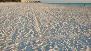 A wide sandy beach with footprints leading towards the water, a clear sky, and buildings in the background. - Westwinds, paradise and heaven all rolled into one (Treasure Island)