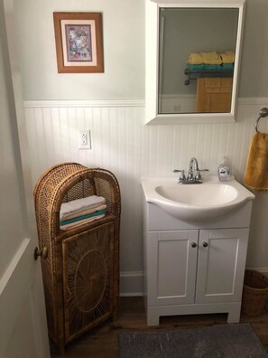 A bathroom with a white vanity, a wicker basket with towels, a framed picture on the wall, and a towel rack with folded towels.