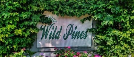 A sign with the text "Wild Pines" surrounded by lush greenery and flowering plants.