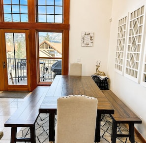 A dining area with a wooden table and benches, large windows with white frames, and a view of the outside.