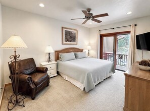 Une chambre avec un lit, une chaise, des tables de chevet, un ventilateur de plafond, un tableau au mur et un balcon avec vue.