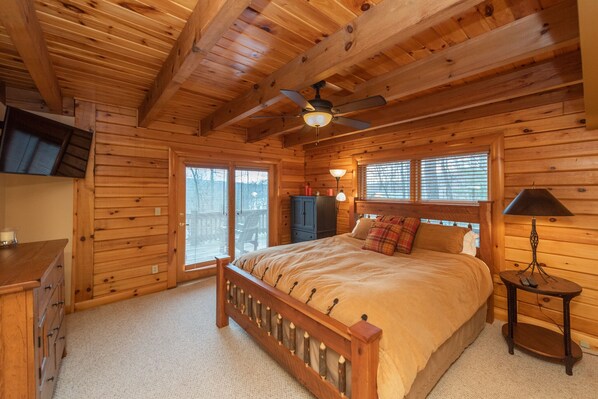 Ein Schlafzimmer in einer Blockhütte mit einem hölzernen Bett, einem Deckenventilator, einem Nachttisch und einem an der Wand befestigten Fernseher.