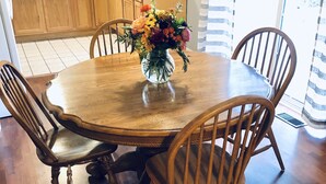 Uma área de jantar com mesa e cadeiras de madeira, um vaso de flores e uma cozinha ao fundo.