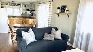 Sala de estar com sofá cinza escuro, mesa de jantar de madeira e cadeiras. Cozinha com armários brancos e janela visível ao fundo.