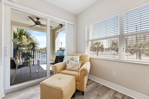 A room with a beige armchair, a matching ottoman, a sliding glass door leading to an outdoor area with a patio, and a large window with blinds.