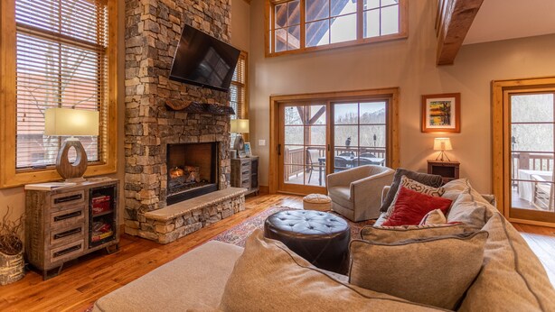 A cozy living room with a stone fireplace, comfortable seating, and large windows offering views of the outdoors. - Bag End at Eagles Nest - Game room, great location in the Villages of Eagles Nest + many amenities (Banner Elk)