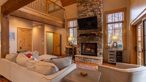 A spacious living room with a stone fireplace, a high wooden ceiling, and a large window with blinds. - Bag End at Eagles Nest - Game room, great location in the Villages of Eagles Nest + many amenities (Banner Elk)