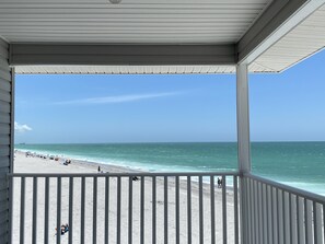 Strandudsigt fra en overdækket balkon med gelænder, med udsigt over havet og badegæster.