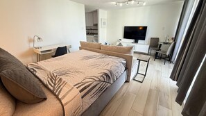 A modern hotel room with a bed, a desk, a TV, and a view of the kitchen.