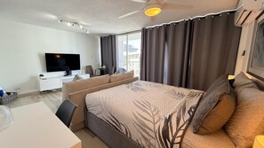 A hotel room with a bed, a TV, a desk, and a window with curtains. - CB118-St Balcony & Ocean View (Carolina)