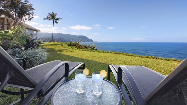 A table with two glasses of water and a lemon slice, overlooking a grassy area with a view of the ocean and a mountain range. - Panoramic Breathtaking Ocean-Sunset-Bali Hai Views A/C + Surf Crashing Below! (Princeville)