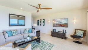 A modern living room with a sofa, coffee table, and a chair. There is a ceiling fan, a painting, and a lamp on the wall. - Panoramic Breathtaking Ocean-Sunset-Bali Hai Views A/C + Surf Crashing Below! (Princeville)