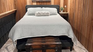 Una camera da letto con una testiera in legno, un letto con copripiumino grigio, un cuscino decorativo, un comodino con una lampada e un quadro appeso al muro.