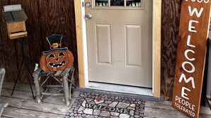 Una porta di legno con un pannello in vetro smerigliato, un tappetino d'ingresso con un motivo a tema Halloween, un piccolo sgabello con una zucca intagliata e un cartello con la scritta "Benvenuti cani, persone tollerate".