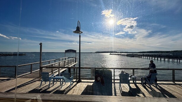 A pier with chairs and a bench, a sign reading 'Demon Bay Beach,' and a view of the water and distant land.