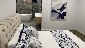Un dormitorio moderno con cabecera capitonada, una cama grande con ropa de cama floral, un baño con bañera y una obra de arte colgada en la pared.