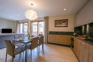 A modern kitchen with wooden cabinets, a dining area with a table set for two, and a living area with a sofa and TV.