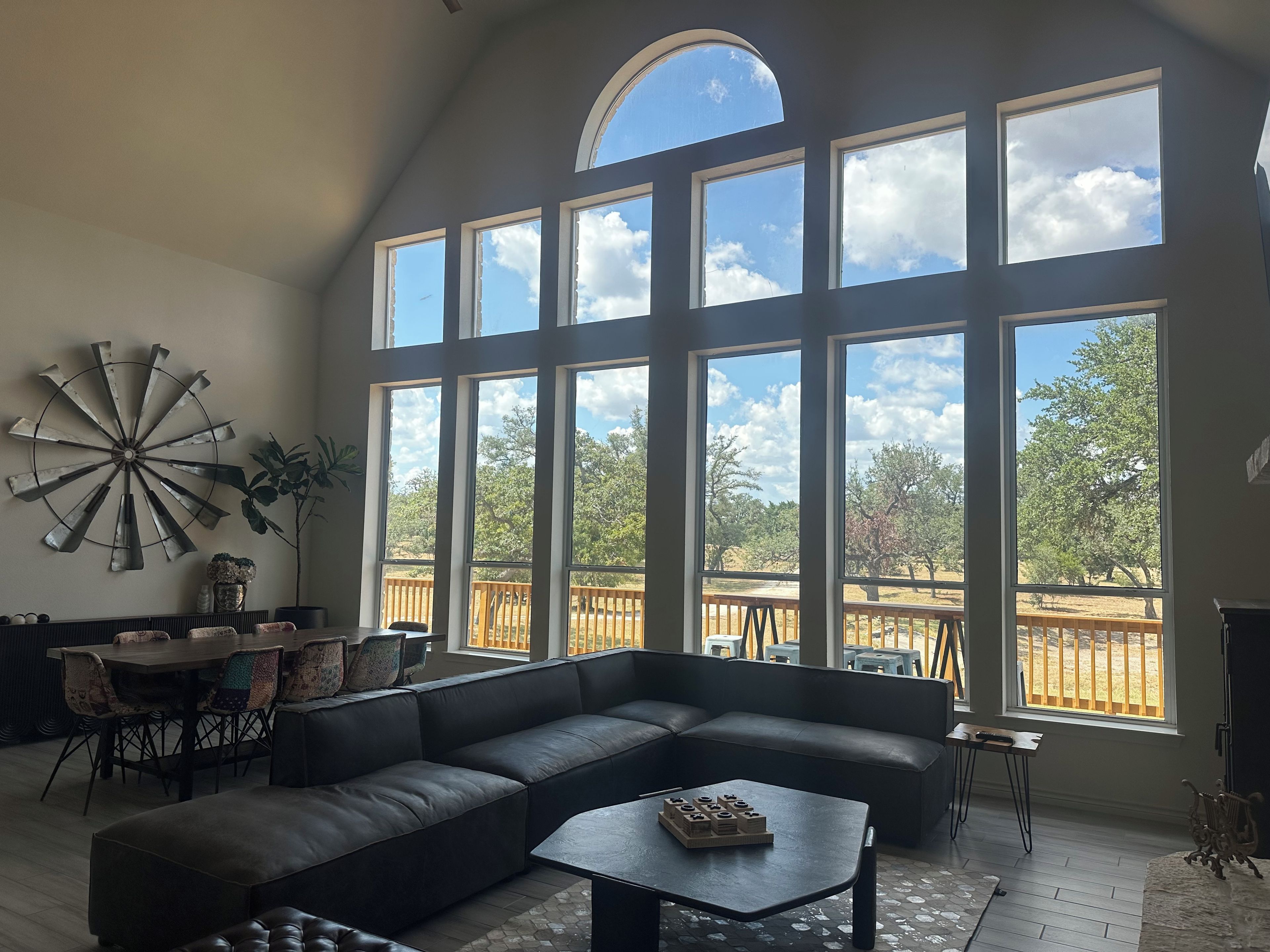 A spacious living room with a large sectional sofa, a dining area with chairs, and a large window with a view of trees and a wooden deck.