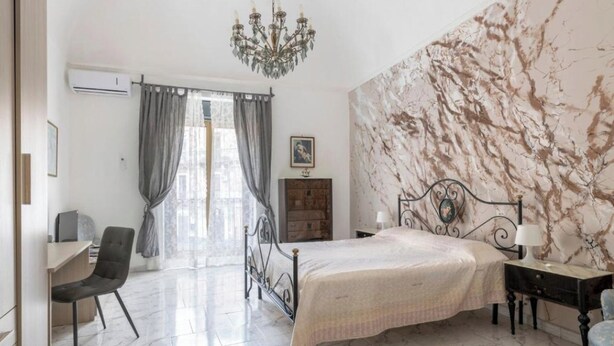 A bedroom with a bed, a desk, a chair, a chandelier, and a marble-patterned wall.