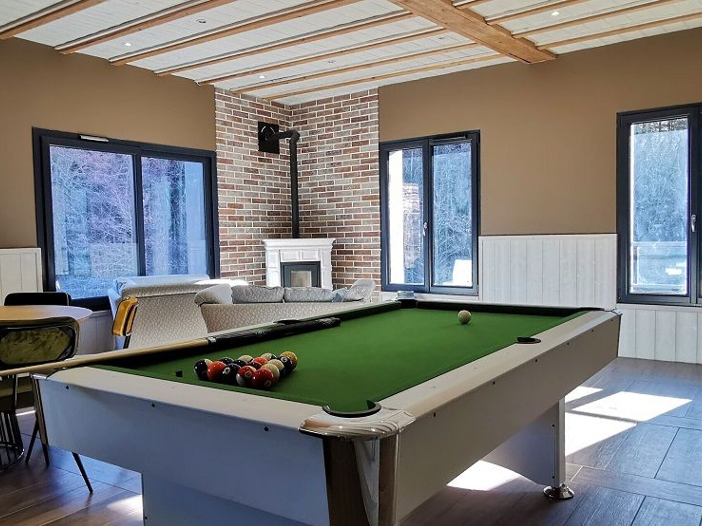 Une table de billard avec des billes, un canapé et une cheminée dans une pièce dotée de grandes fenêtres et d’un mur en briques.