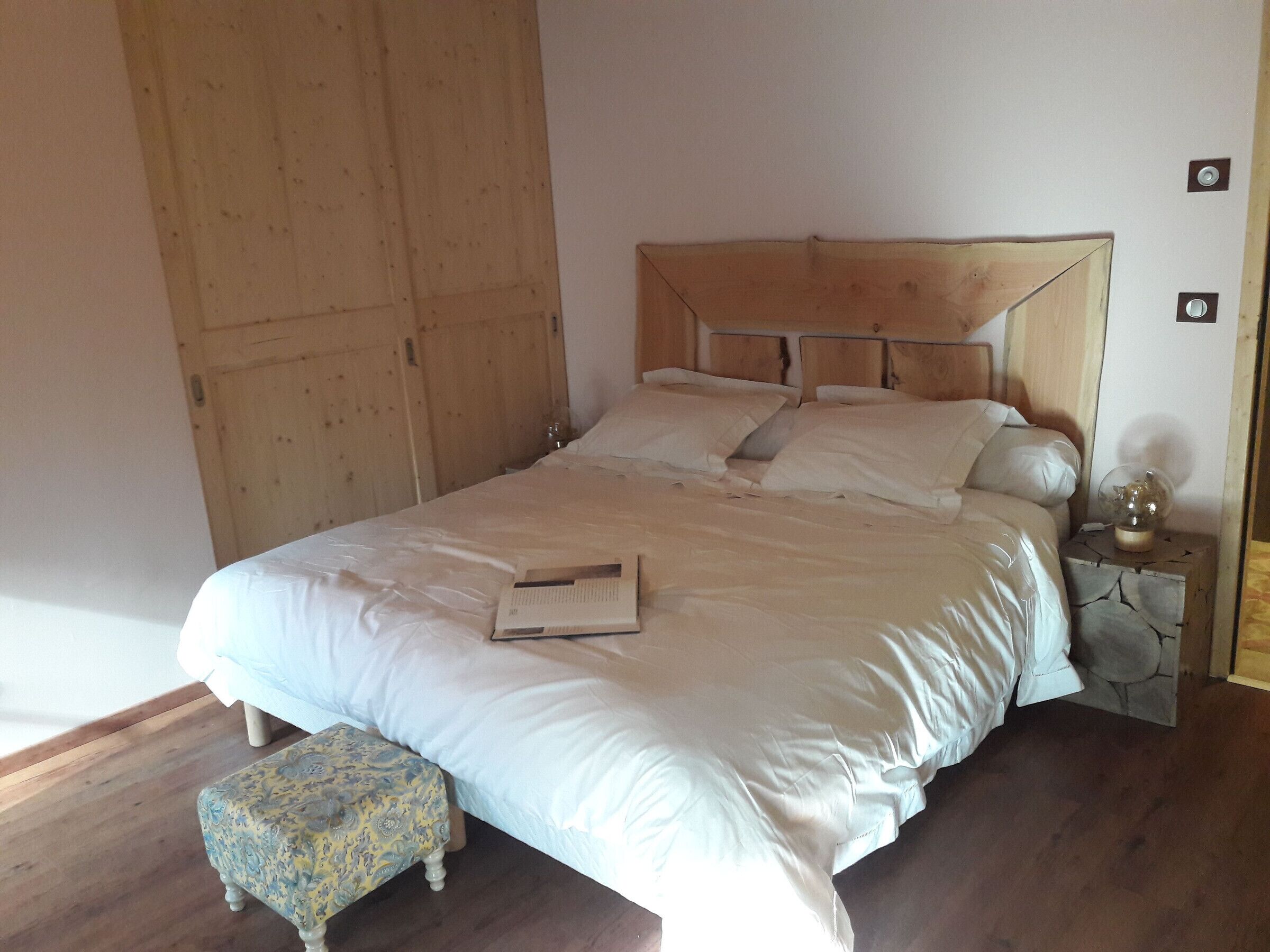 A bedroom with a wooden headboard, a bed with white bedding, a small bedside table with a lamp, and a patterned ottoman.