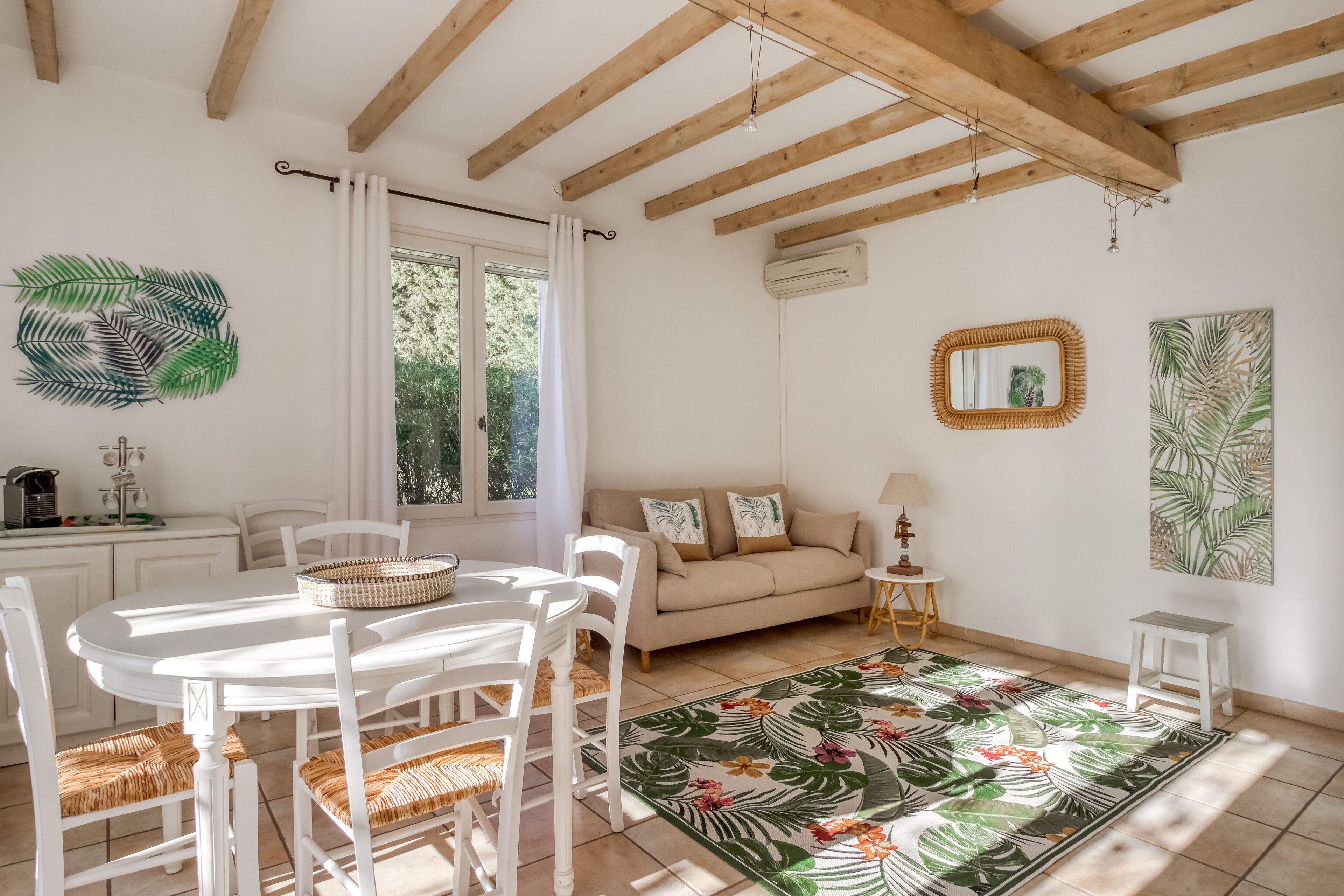 Une pièce lumineuse et bien éclairée, avec un plafond en bois, une table à manger blanche avec des chaises, un canapé et un tapis aux motifs tropicaux.