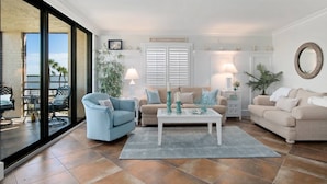 Ein Wohnzimmer mit einer großen Schiebeglaswand, die auf einen Balkon führt, einem beigen Sofa, einem blauen Sessel, einem weißen Couchtisch und einem kleinen Beistelltisch mit einer Lampe.
