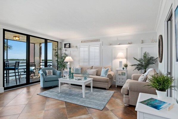 A spacious living room with a large sliding glass door leading to a balcony, a beige sofa, a blue armchair, and a white coffee table.