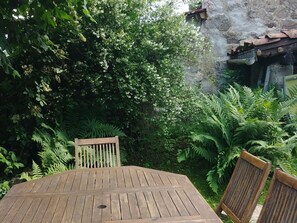 Mesa y sillas de madera en un jardín exuberante, con follaje denso y un muro de piedra al fondo.