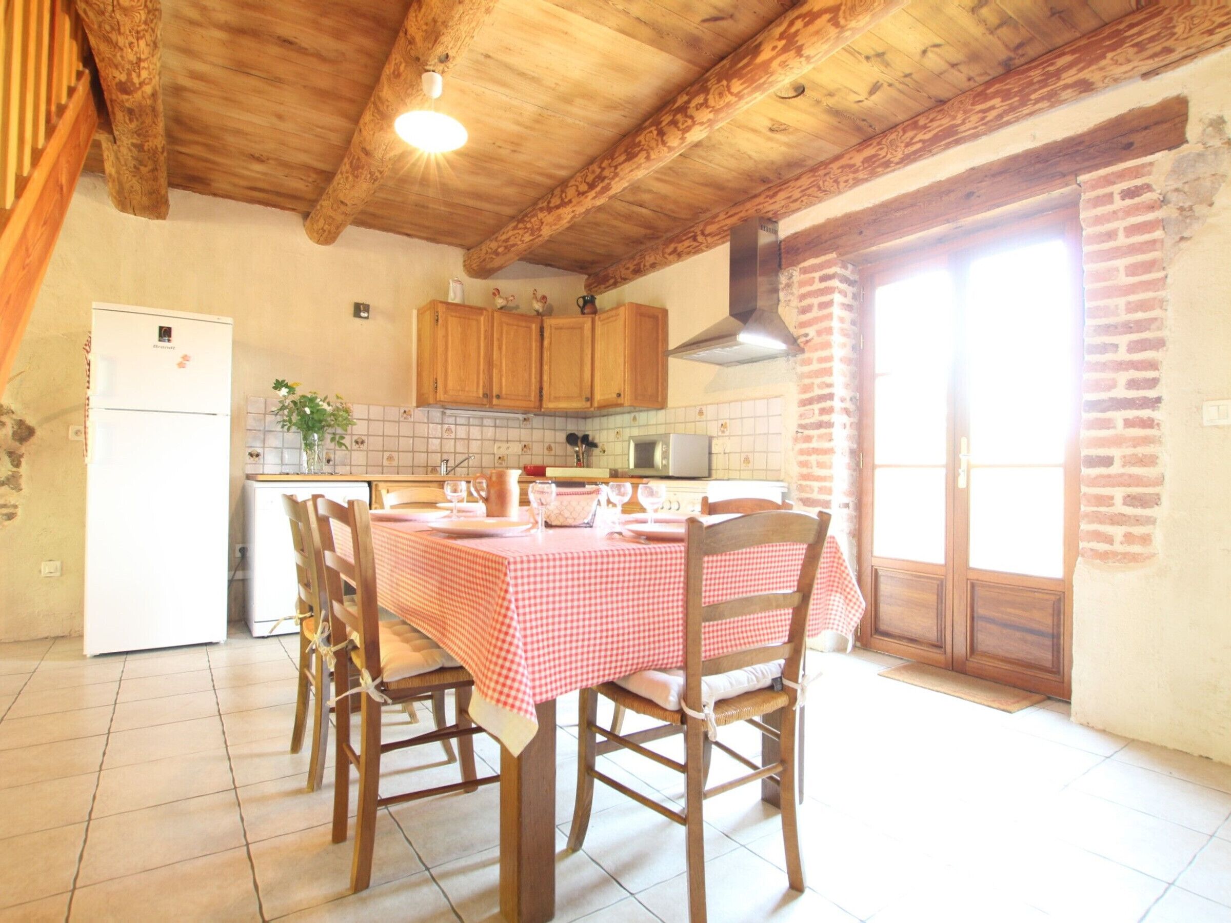 Une cuisine avec des meubles de rangement en bois, un pare-feu en carrelage, une table à manger recouverte d’une nappe à carreaux et un réfrigérateur.