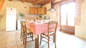 A kitchen with wooden cabinets, a tiled backsplash, a dining table with a checkered tablecloth, and a refrigerator. - Charming 19th-Century Farmhouse near Loire Gorges, Cozy Interior, Baby Equipment, Optional Services (Aurec-sur-Loire)