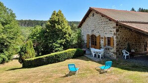 Ein Gebäude aus Stein mit einem Ziegeldach, einer kleinen Terrasse mit weißen Stühlen und einem gepflegten Rasen.