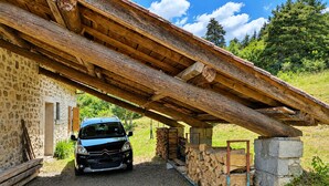 Ein Auto, das unter einer hölzernen Struktur abgestellt ist, mit einem Steinhaus im Hintergrund.