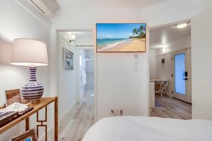 A hotel room with a bed, a desk with a lamp, a painting of a beach, and a view of the beach through an open door.