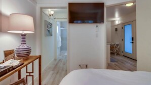 Ein Hotelzimmer mit Bett, Nachttisch mit Lampe, Wand-Fernseher und Blick auf den Flur und ein weiteres Zimmer.