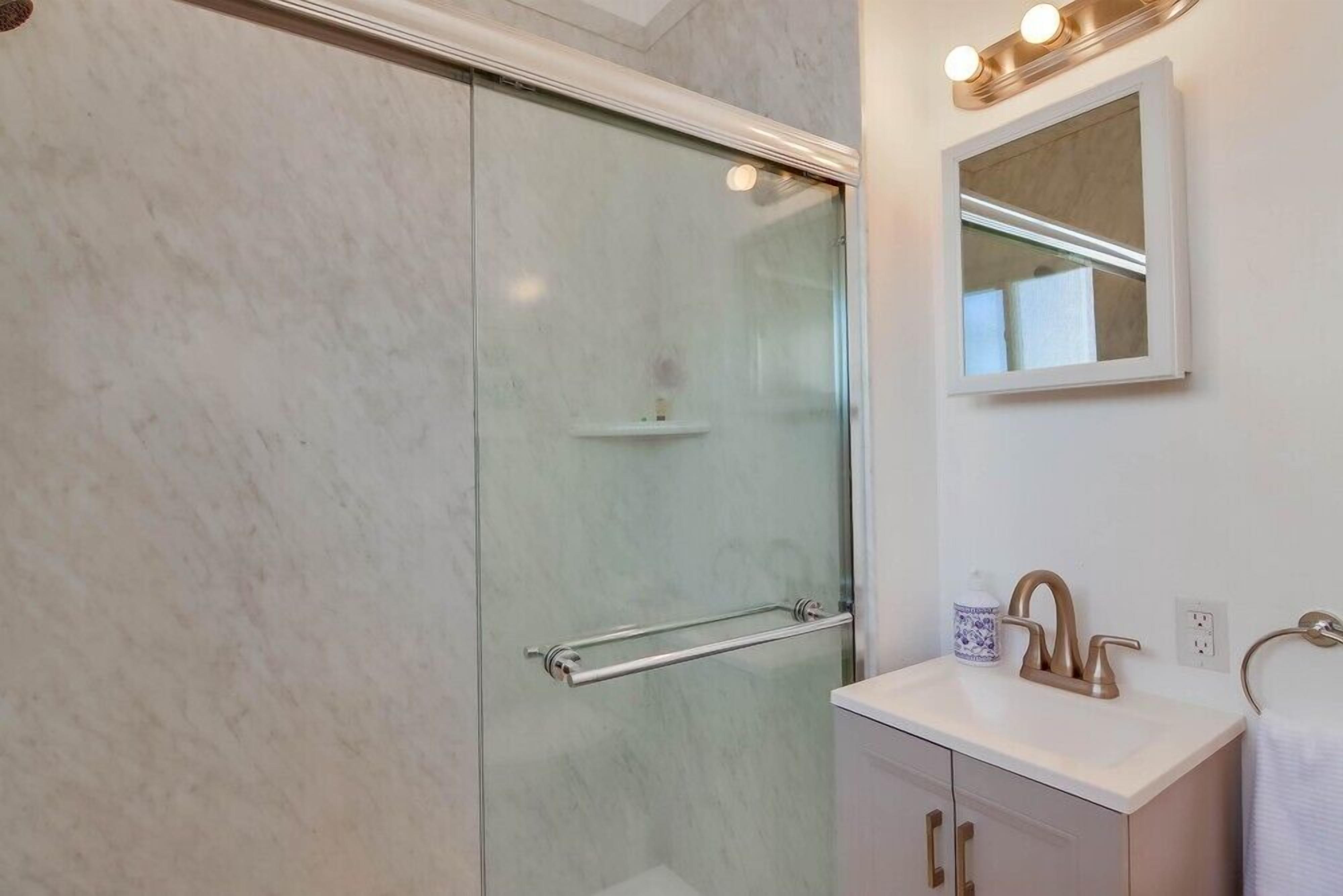 Ein modernes Badezimmer mit einer Glasdusche, einem weißen Waschbecken mit Goldarmatur und einem Spiegel mit Beleuchtung darüber.