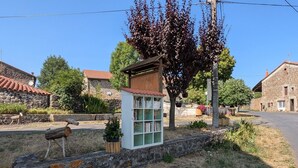 Un paysage rural avec des bâtiments en pierre, une petite structure en bois remplie de livres et un arbre aux feuilles rouges.