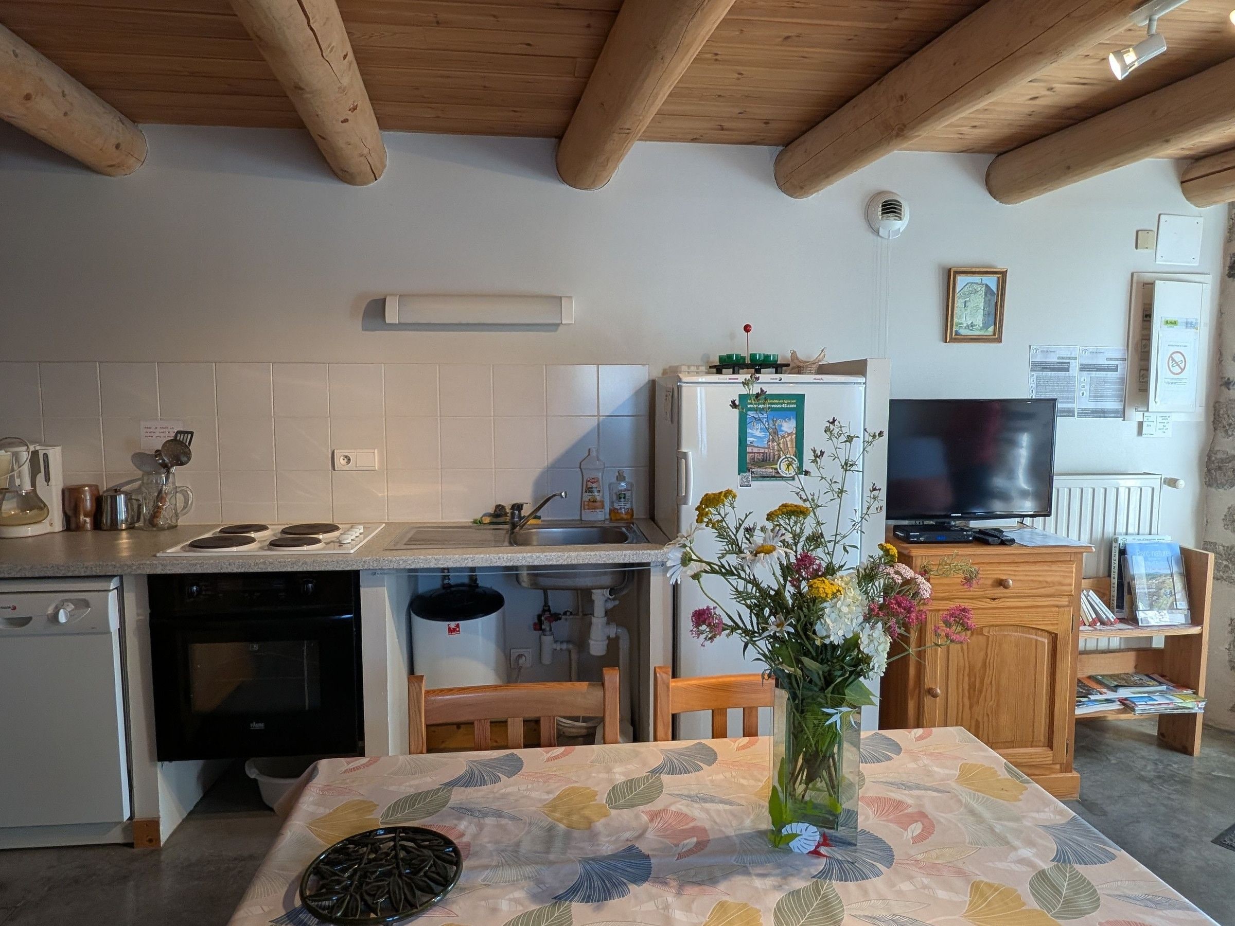 Une cuisine avec des poutres en bois, un réfrigérateur, une cuisinière et une table à manger recouverte d’une nappe à motifs floraux.