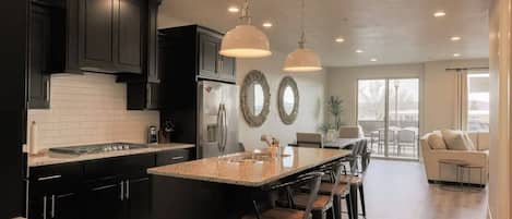 A modern kitchen with dark cabinets, a central island with bar stools, pendant lighting, and a view to a living area with a sofa and a table.