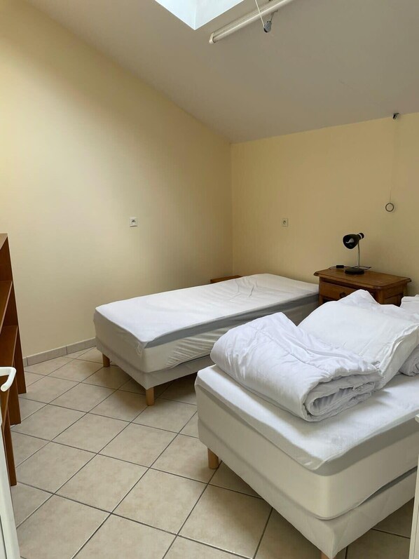 A small, clean room with two single beds, a bedside table with a lamp, and a skylight.